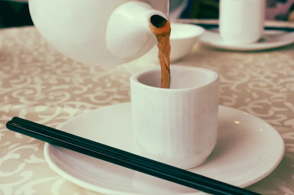 Close-up shot at a slight side angle of Chinese tea being poured from a white ceramic teapot into a porcelain teacup resting on a saucer with chopsticks, capturing a traditional yum cha tea ritual in an elegant dining setting.
