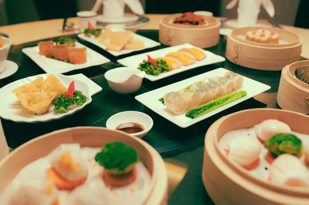 Wide-angle, slightly elevated shot of a large round dining table set for dim sum at a Chinese restaurant in Singapore, featuring an array of white rectangular plates and bamboo steamers holding translucent shrimp dumplings, fried pastries, sliced rolls, and garnished vegetables, with small porcelain bowls of dipping sauces arranged between dishes. The composition is centered and symmetrical, with warm ambient indoor lighting highlighting glossy ceramic surfaces, wooden steamers, and the refined presentation of Cantonese dim sum.