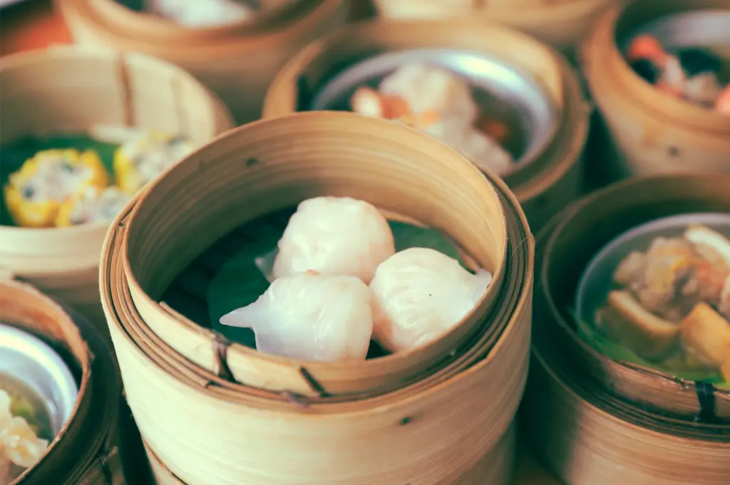 Close-up shot at a slight downward angle of fresh dim sum har gow shrimp dumplings served in traditional bamboo steamers, with assorted dumplings in the background, highlighting authentic Cantonese cuisine in Singapore.