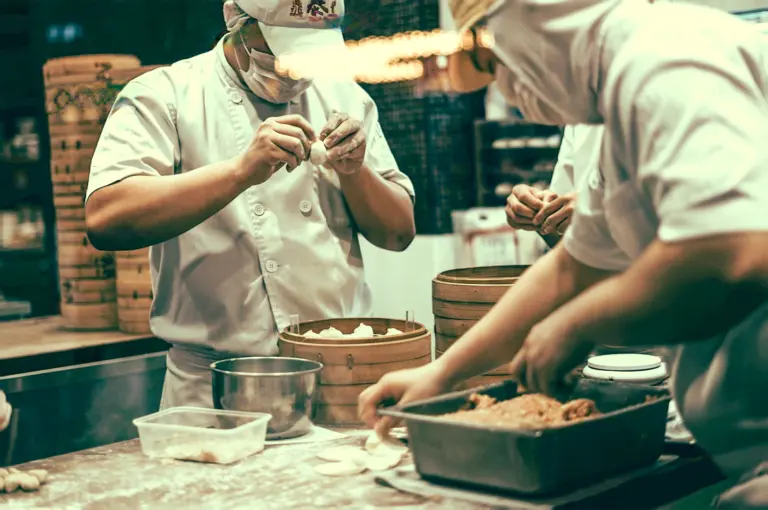 Professional kitchen scene showing chefs hand-making fresh dim sum dumplings and placing them into bamboo steamers on a stainless steel worktable, captured at a medium eye-level angle.