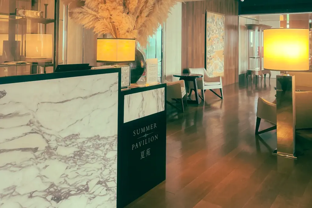 Low-angle wide shot of the Summer Pavilion restaurant reception area at The Ritz-Carlton, featuring a marble host stand, warm ambient lighting, pampas grass floral arrangement, and modern wooden interiors.