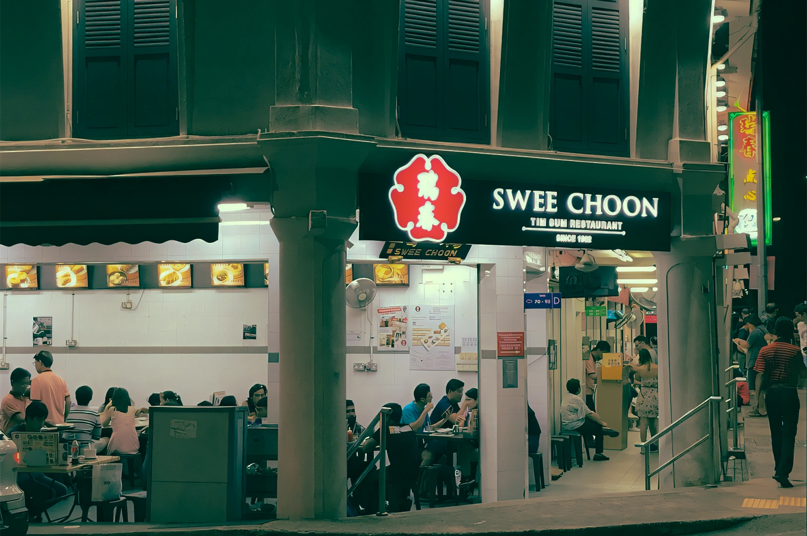 Wide-angle street-level shot of Swee Choon Tim Sum Restaurant along Jalan Besar at night, showing the illuminated storefront, indoor dining area, and bustling late-night dim sum crowd in Singapore.