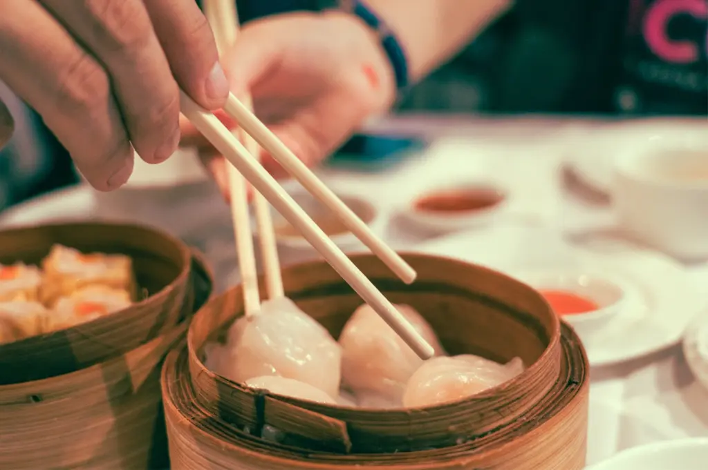 Close-up, shallow-depth-of-field shot of two hands holding chopsticks, each lifting har gow shrimp dumplings from bamboo steamer baskets on a restaurant table during traditional Cantonese dim sum service.