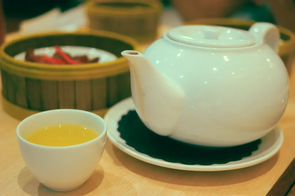 Close-up side-angle shot of a white ceramic teapot and small porcelain tea cup filled with amber Chinese tea on a wooden table, capturing the quiet beginning of a traditional yum cha tea ritual.