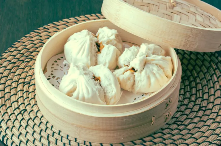 Top-down food photography of steamed char siu bao, fluffy Chinese BBQ pork buns served in a bamboo steamer on a woven placemat. Shot angle: Overhead / top-down
