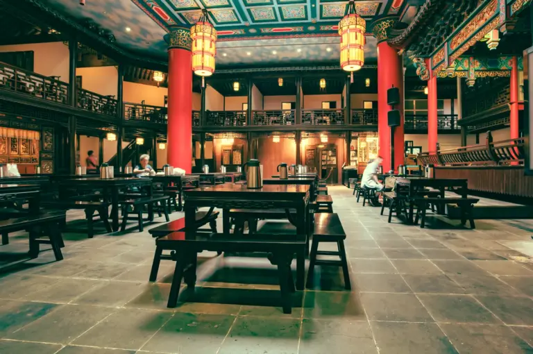 Wide-angle interior architecture shot of a traditional Chinese banquet hall with red columns, hanging lanterns, wooden tables, and an open central seating layout.