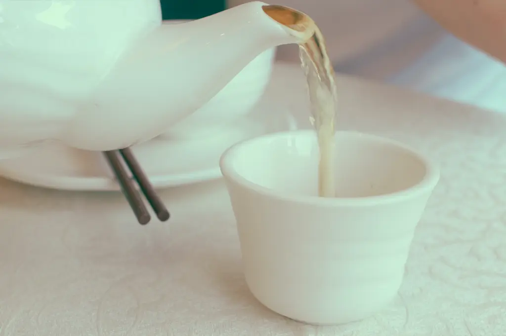 Close-up angled shot of hot Chinese tea pouring from a white ceramic teapot into a handle-less porcelain cup, highlighting the first pour ritual central to the yum cha experience.