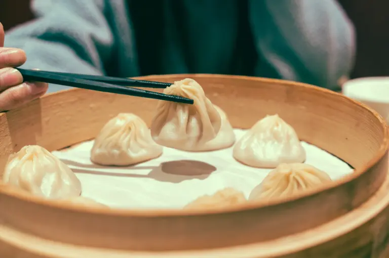 Eye-level close-up of xiao long bao soup dumplings in a bamboo steamer, with chopsticks lifting one dumpling to show its delicate pleats. Shot angle: Eye-level close-up