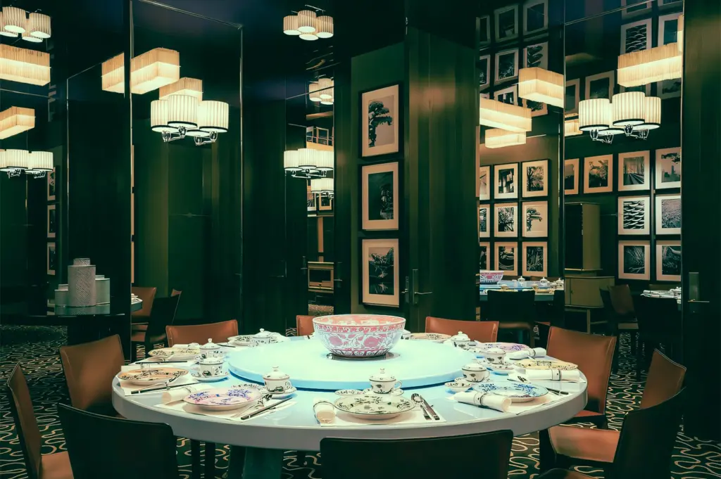 Eye-level wide-angle shot of a private dining room at Summer Pavilion, The Ritz-Carlton, showcasing a round banquet table with lazy Susan, luxurious tableware, mirrored walls, framed artwork, and elegant pendant lighting.