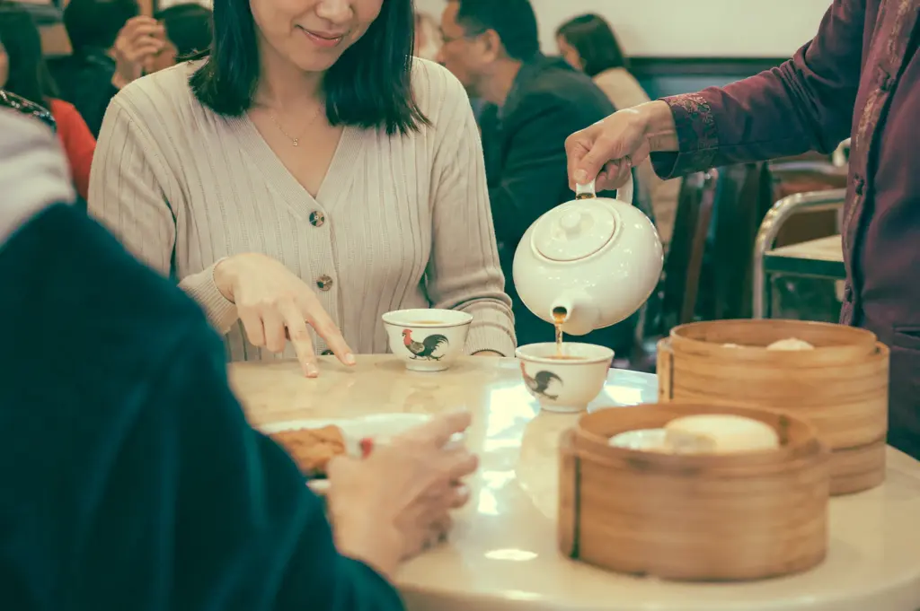Medium close-up shot of diners as one person pours tea while another performs the finger tap gesture on the tabletop, expressing silent thanks in traditional dim sum etiquette.