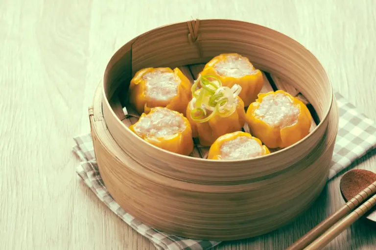 Angled tabletop shot of traditional siu mai dumplings with pork and shrimp, neatly arranged in a bamboo steamer and garnished with scallions. Shot angle: Three-quarter / angled top-down