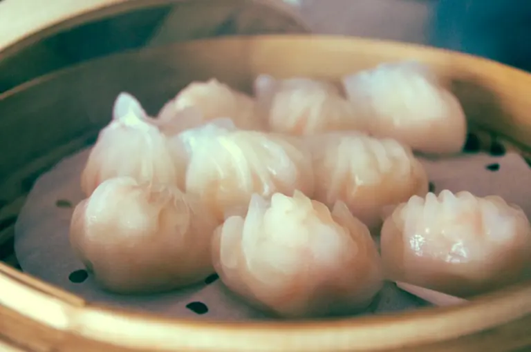 Close-up food shot of translucent har gow shrimp dumplings steaming in a bamboo basket, highlighting the glossy rice wrappers.