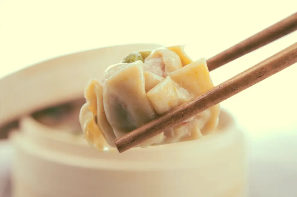 Extreme close-up shot of a single dim sum dumpling held gently between chopsticks above a bowl, symbolizing mindful sharing and respect for the last piece in yum cha etiquette.