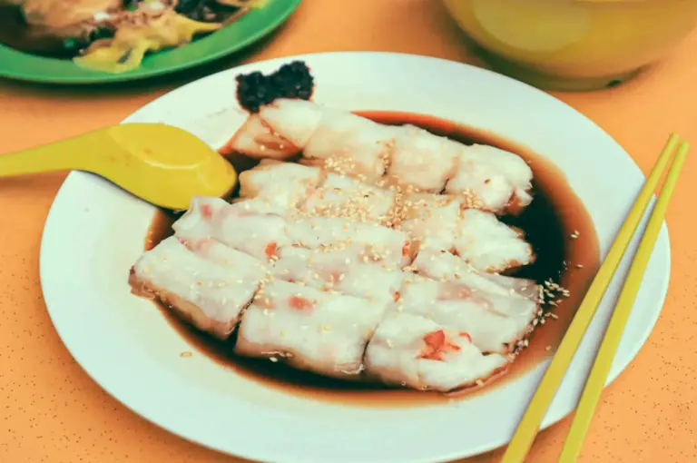 Top-down view of cheong fun rice noodle rolls served with soy sauce and sesame seeds, photographed on a restaurant table with chopsticks.