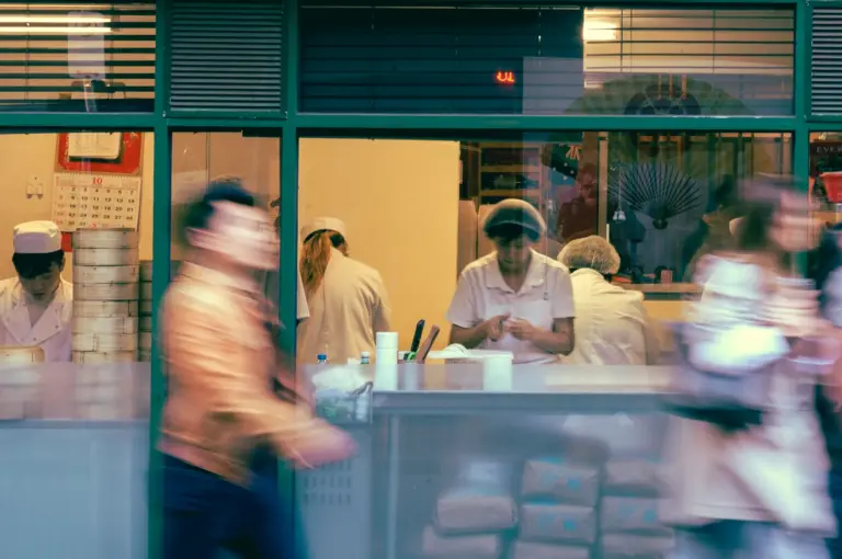 Wide-angle street-level shot of a dim sum kitchen behind glass with chefs preparing dumplings while blurred pedestrians pass by, capturing the fast-paced atmosphere of an urban dim sum restaurant.