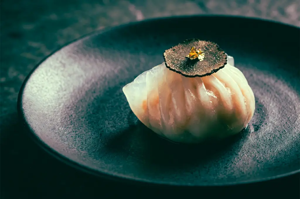 A single, elegant Har Gow (steamed shrimp dumpling) rests on a textured, dark charcoal-ceramic plate. The dumpling features a translucent, pleated skin through which the pink shrimp filling is visible.
