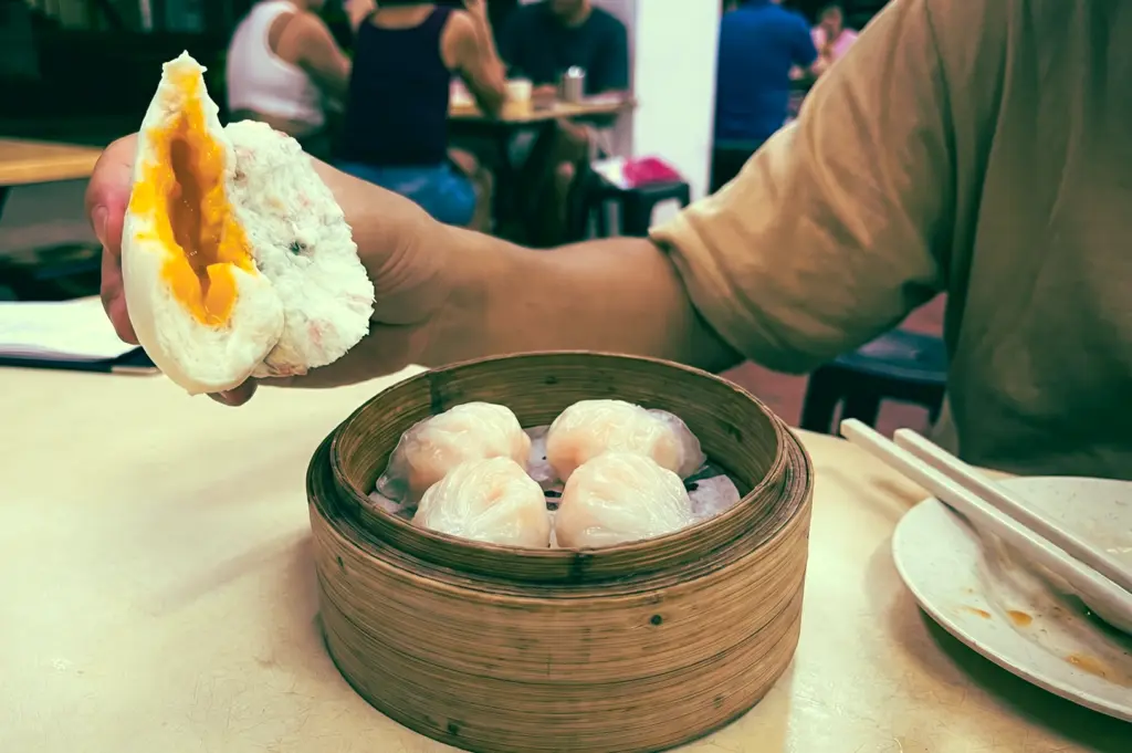 Close-up eye-level shot of a salted egg lava bun bursting with golden filling held above a bamboo steamer of har gow dumplings in a late-night dim sum restaurant in Singapore