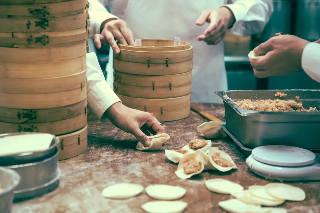Wide-angle slightly high-angle shot of chefs preparing raw dumplings on a floured work surface with stacked bamboo steamers, illustrating traditional dim sum production workflow