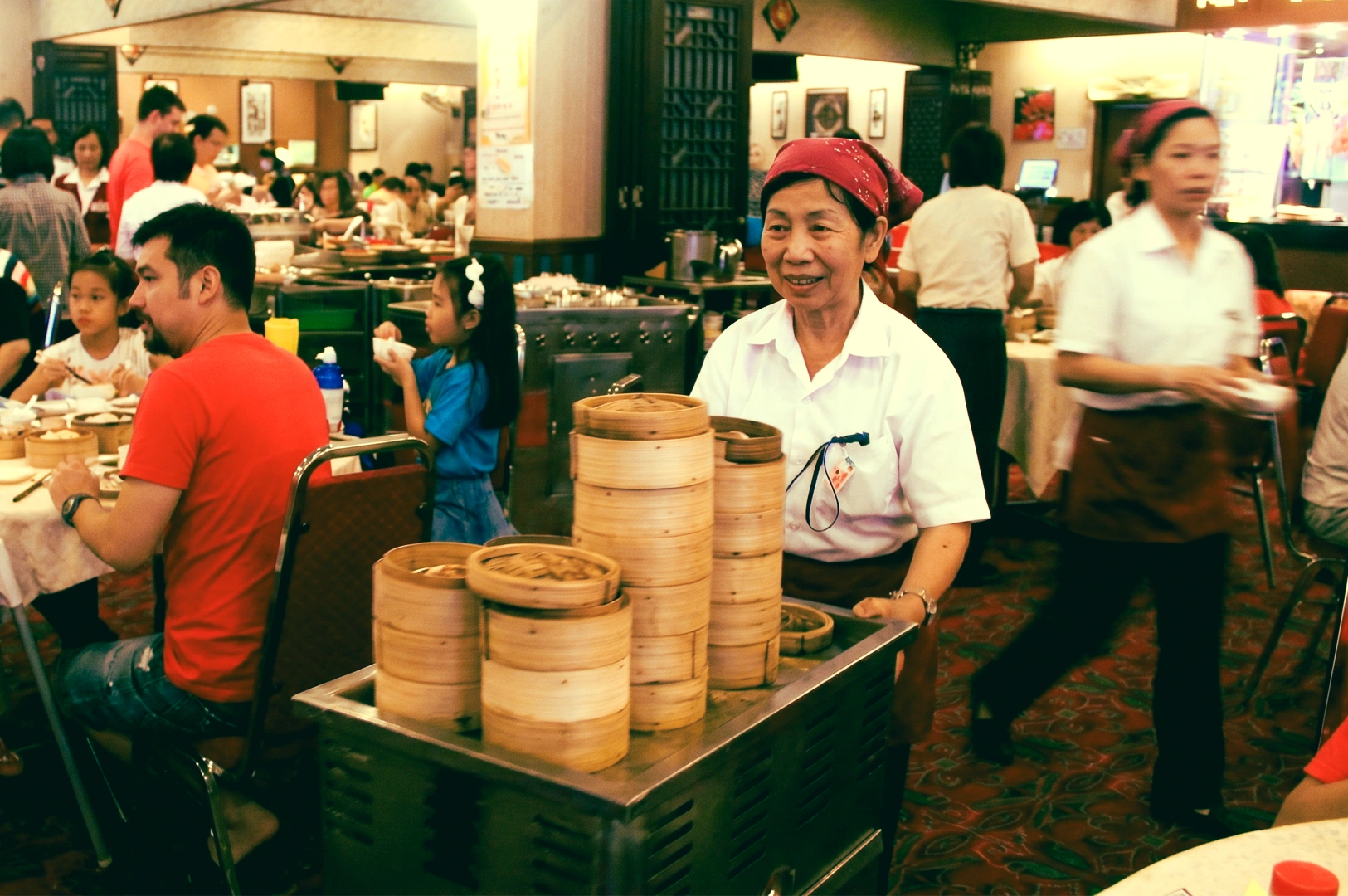 Eye-level candid shot of a busy dim sum restaurant during lunch rush, featuring a server pushing a cart stacked with bamboo steamers through a crowded dining room with diners and waitstaff in motion