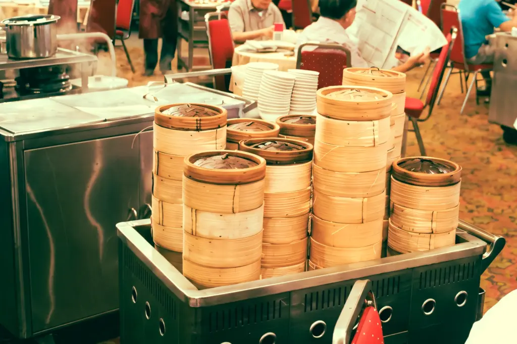 Eye-level shot of a dim sum trolley filled with stacked bamboo steamers beside a restaurant table, showcasing traditional pushcart dim sum service in a busy dining setting.