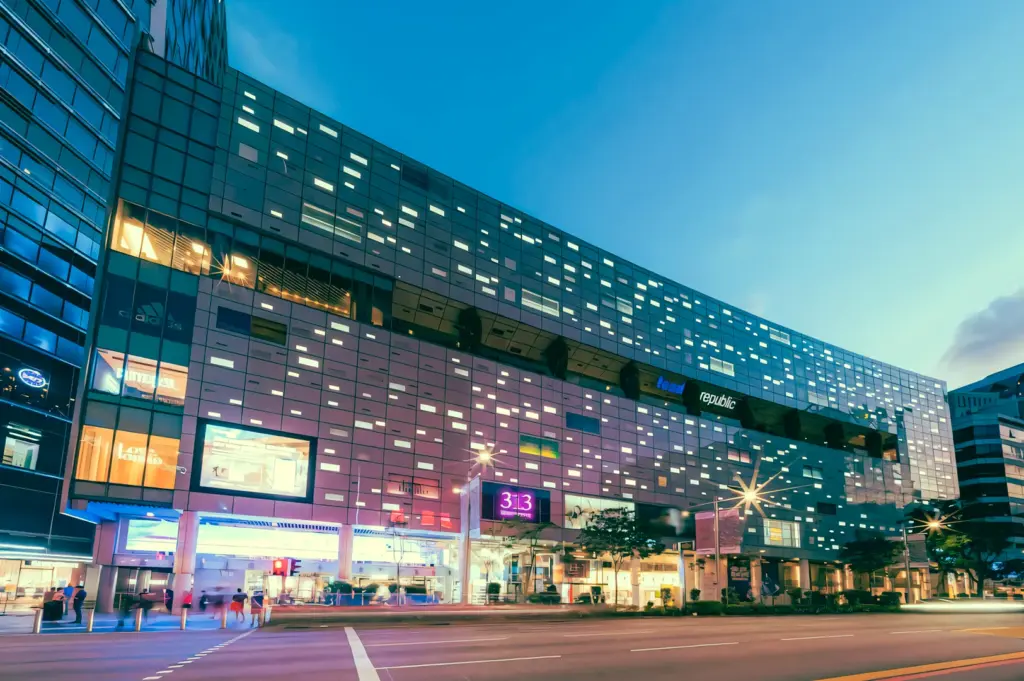 Wide-angle low-angle exterior shot of a modern shopping mall along Orchard Road at dusk, featuring illuminated facade with geometric light patterns, visible storefront signage including ‘313’, and light traffic on the street below
