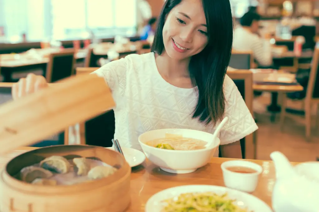 Eye-level medium shot of diner reaching for bamboo steamer while sitting at dim sum table with noodle soup and tea, illustrating modern yum cha dining experience focused on food