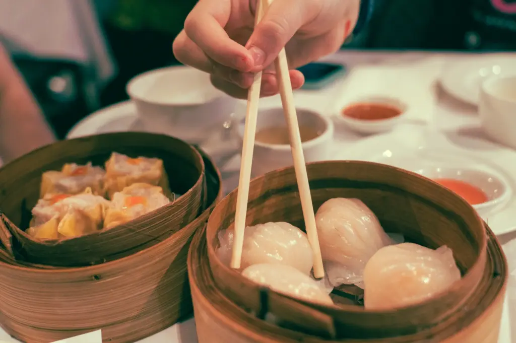 Eye-level close-up shot of chopsticks lifting har gow from a bamboo steamer alongside siew mai dumplings, showcasing freshness and proper dim sum serving presentation