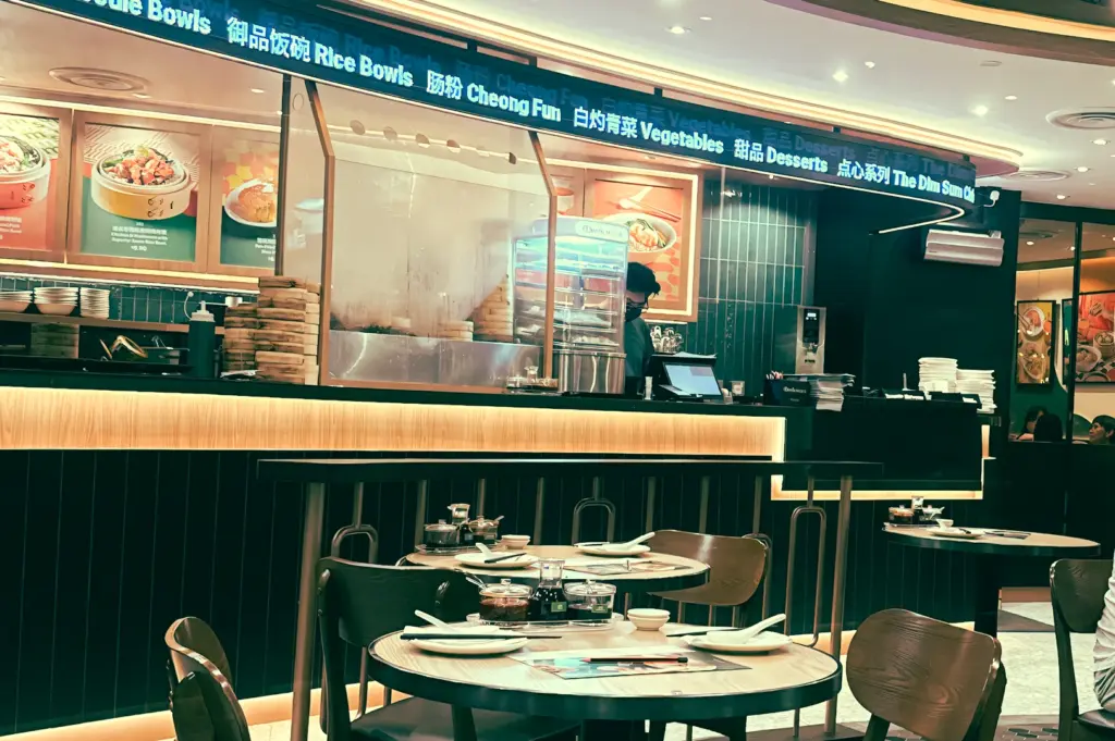 Eye-level wide shot of Dim Sum Club interior counter and dining tables, showing open kitchen, menu boards, and casual mall restaurant layout in Wheelock Place