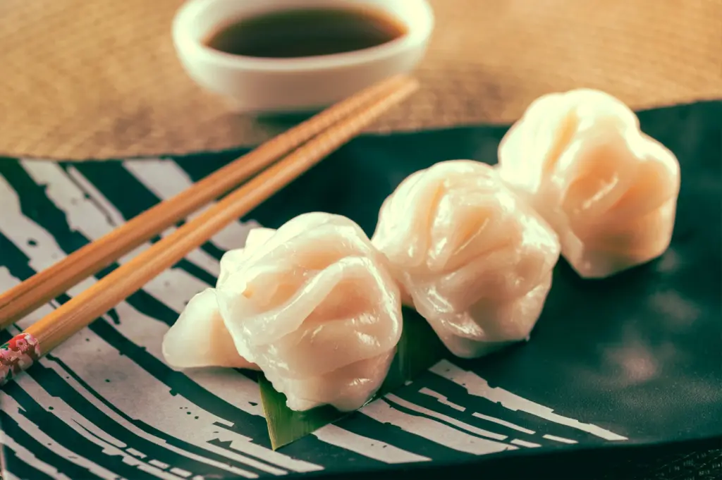 Side-angle close-up shot of har gow shrimp dumplings with translucent wrappers served with chopsticks and dipping sauce, emphasizing delicate pleating and premium dim sum quality