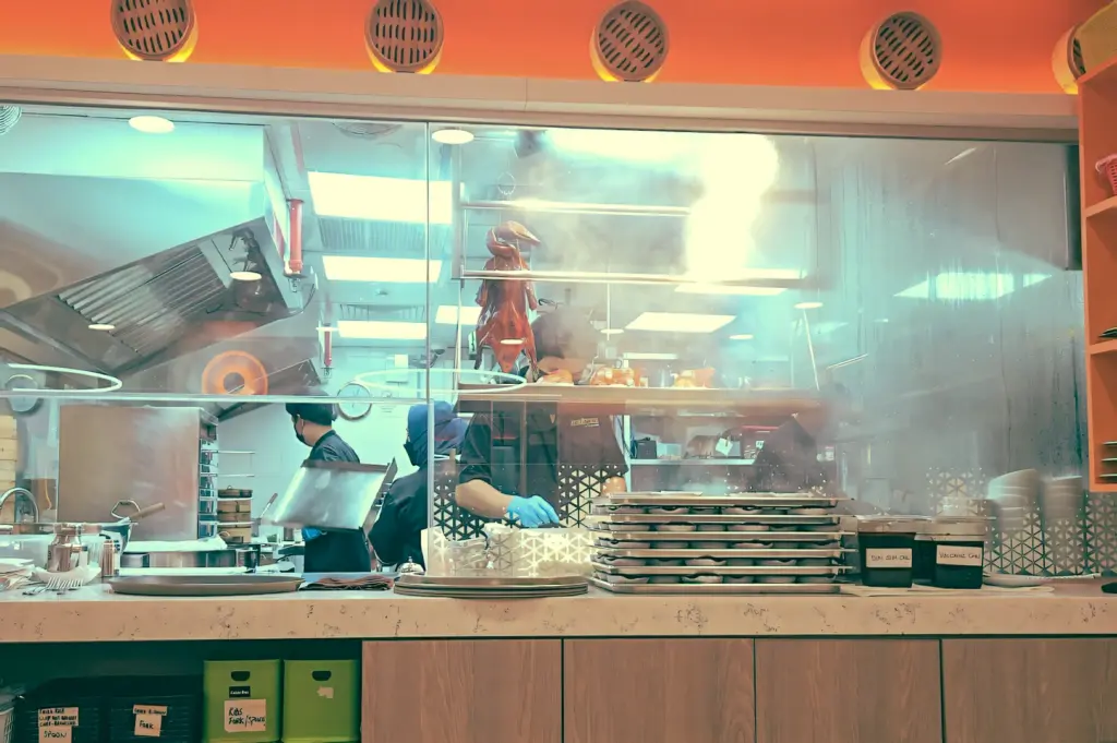 Eye-level wide shot of halal dim sum kitchen at The Dim Sum Place Centrepoint with chefs preparing Cantonese dishes behind glass, steam rising in a busy Orchard Road restaurant