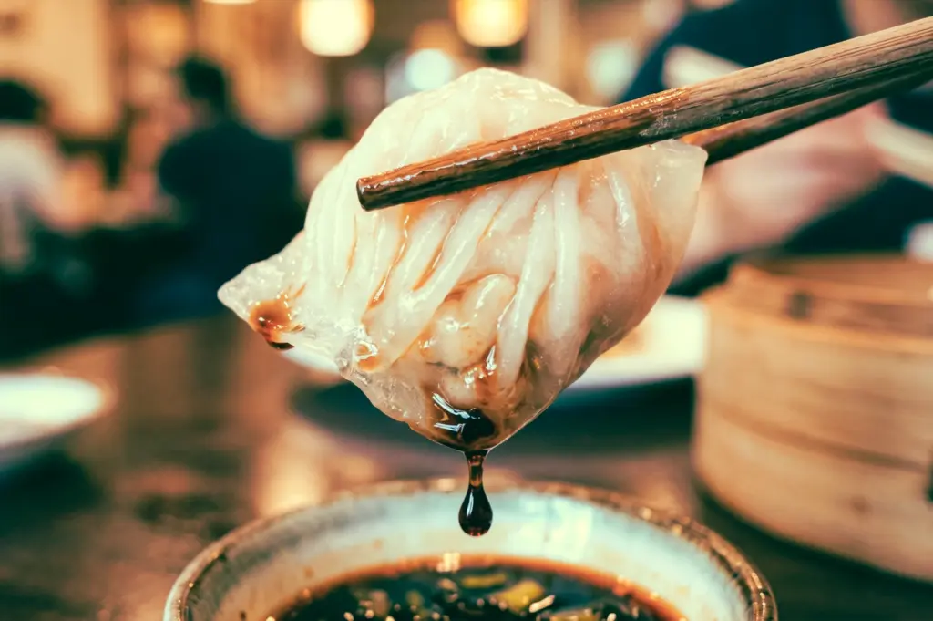 Eye-level macro shot of har gow shrimp dumpling held with chopsticks above dipping sauce, featuring translucent pleated wrapper and dripping soy sauce in a dim sum restaurant setting, highlighting authentic Cantonese yum cha experience