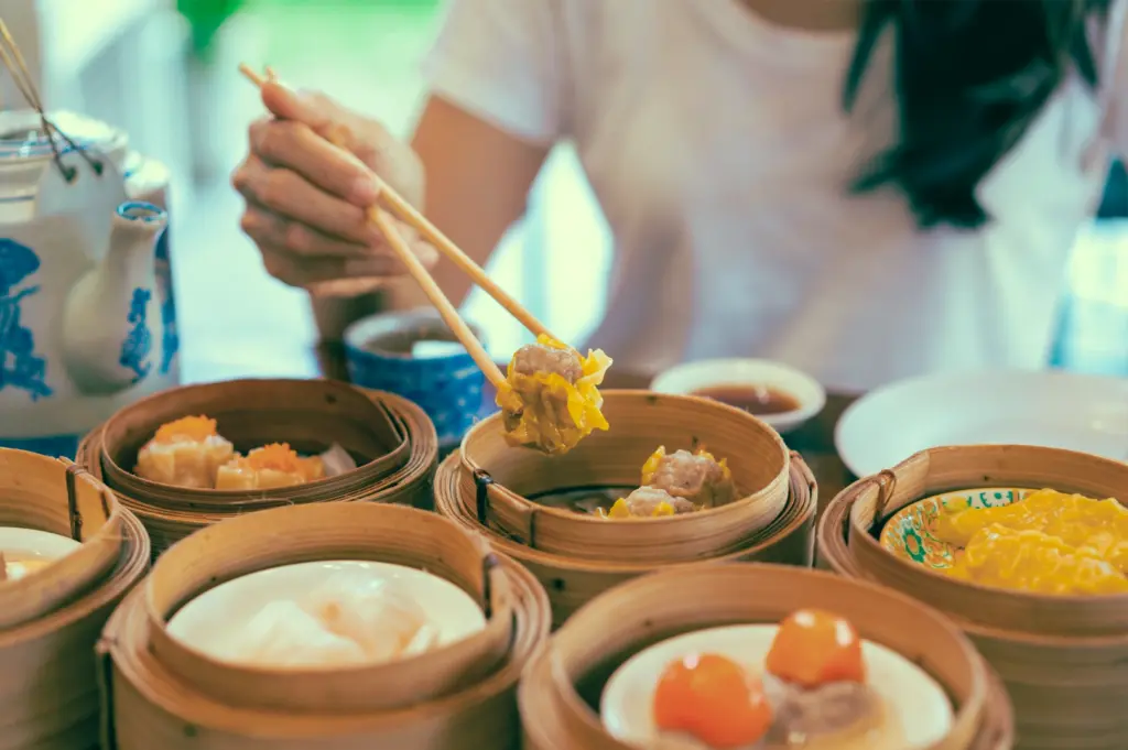 Close-up angled shot of chopsticks lifting siu mai dumpling from bamboo steamer among assorted dim sum, emphasizing variety in yum cha dining