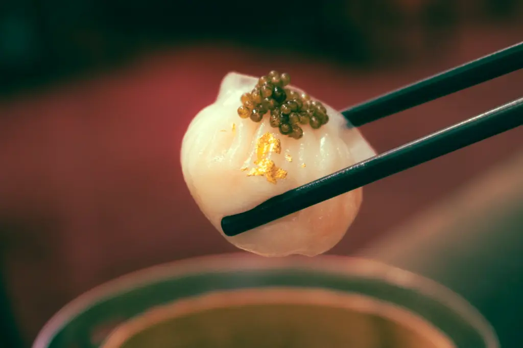 Macro close-up of modern har gow topped with caviar and gold leaf held by chopsticks, showcasing luxury dim sum innovation and translucent dumpling wrapper in contemporary Cantonese cuisine