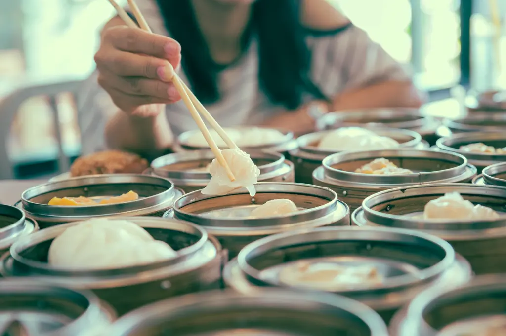Macro close-up of chopsticks picking translucent shrimp dumpling from steaming baskets, showing fluid pacing of dim sum and tea experience