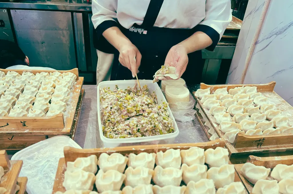 Overhead medium shot of a chef assembling handmade dumplings with fresh pork and vegetable filling, surrounded by trays of raw dim sum in a commercial kitchen showing traditional preparation techniques