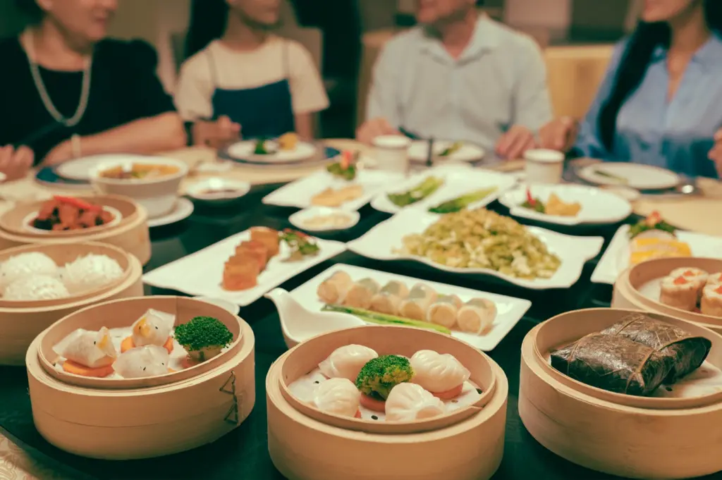 Eye-level wide shot of diners sharing multiple dim sum dishes on a restaurant table, highlighting guest dining experience and peak service period in a dim sum restaurant