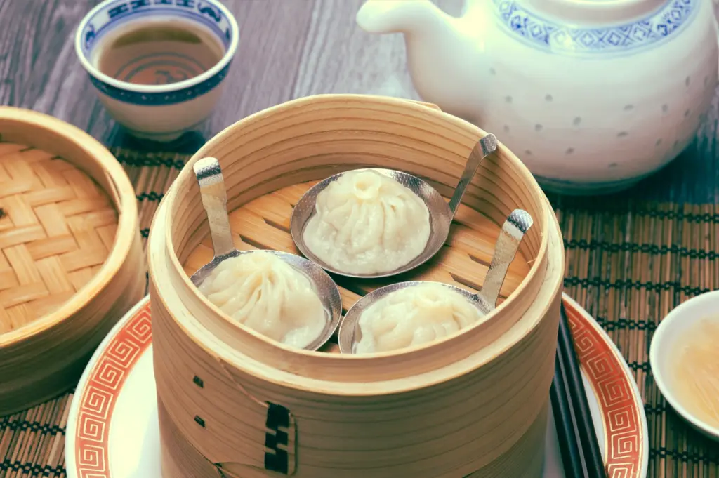 High-angle close-up of xiao long bao in bamboo steamer with tea set nearby, symbolizing balance between dim sum and tea in Chinese dining tradition